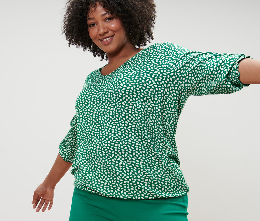Kobieta pozuje w zielonej wzorzystej bluzce i zielonych spodniach stretchowych.