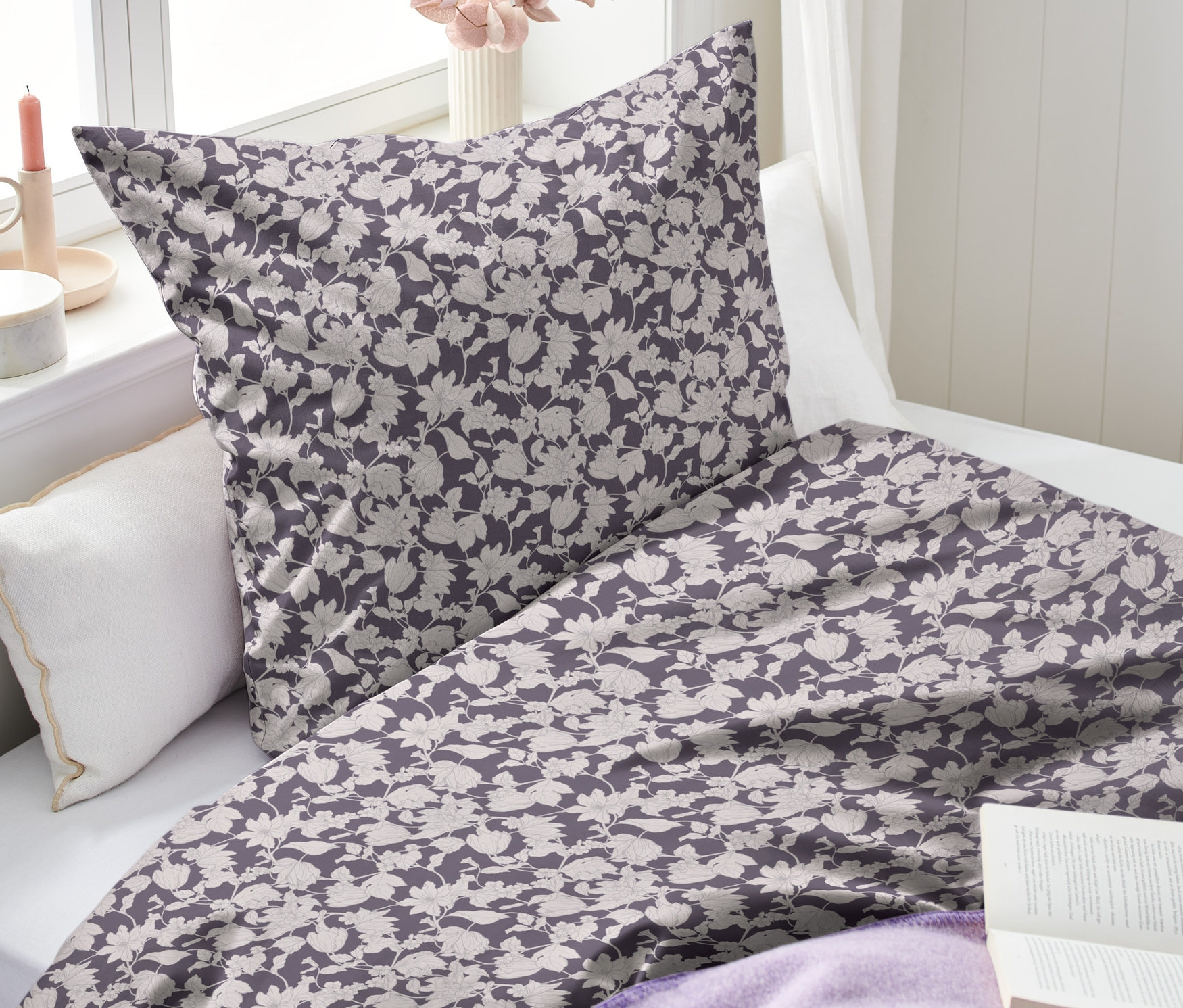Fioletowa pościel z bawełny renforcé w kwiaty leży na łóżku. Obok leży otwarta książka i fioletowy koc.