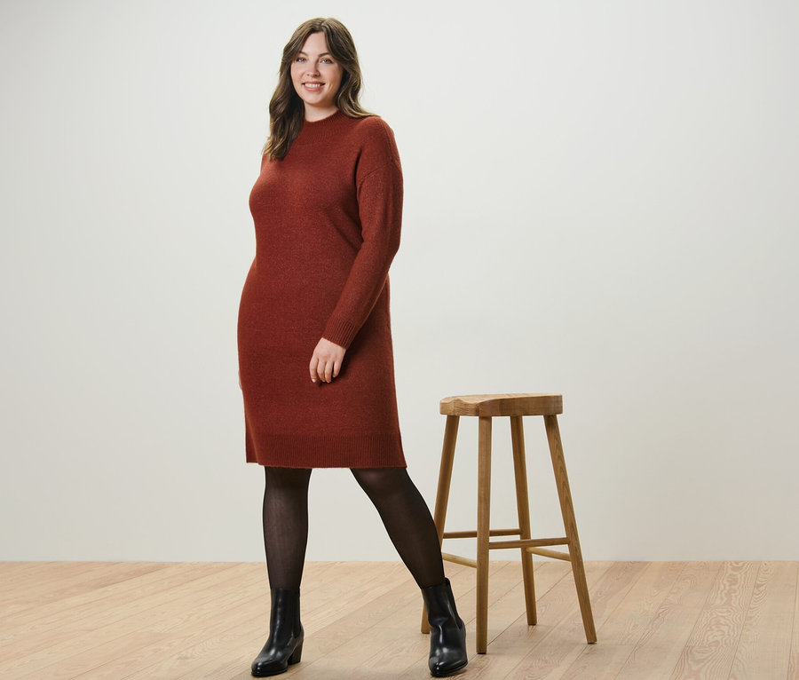 Kobieta pozuje w dzianinowej sukience z zawartością wełny i cienkich rajstopach 50 DEN z efektem przezroczystości obok drewnianego stołka.