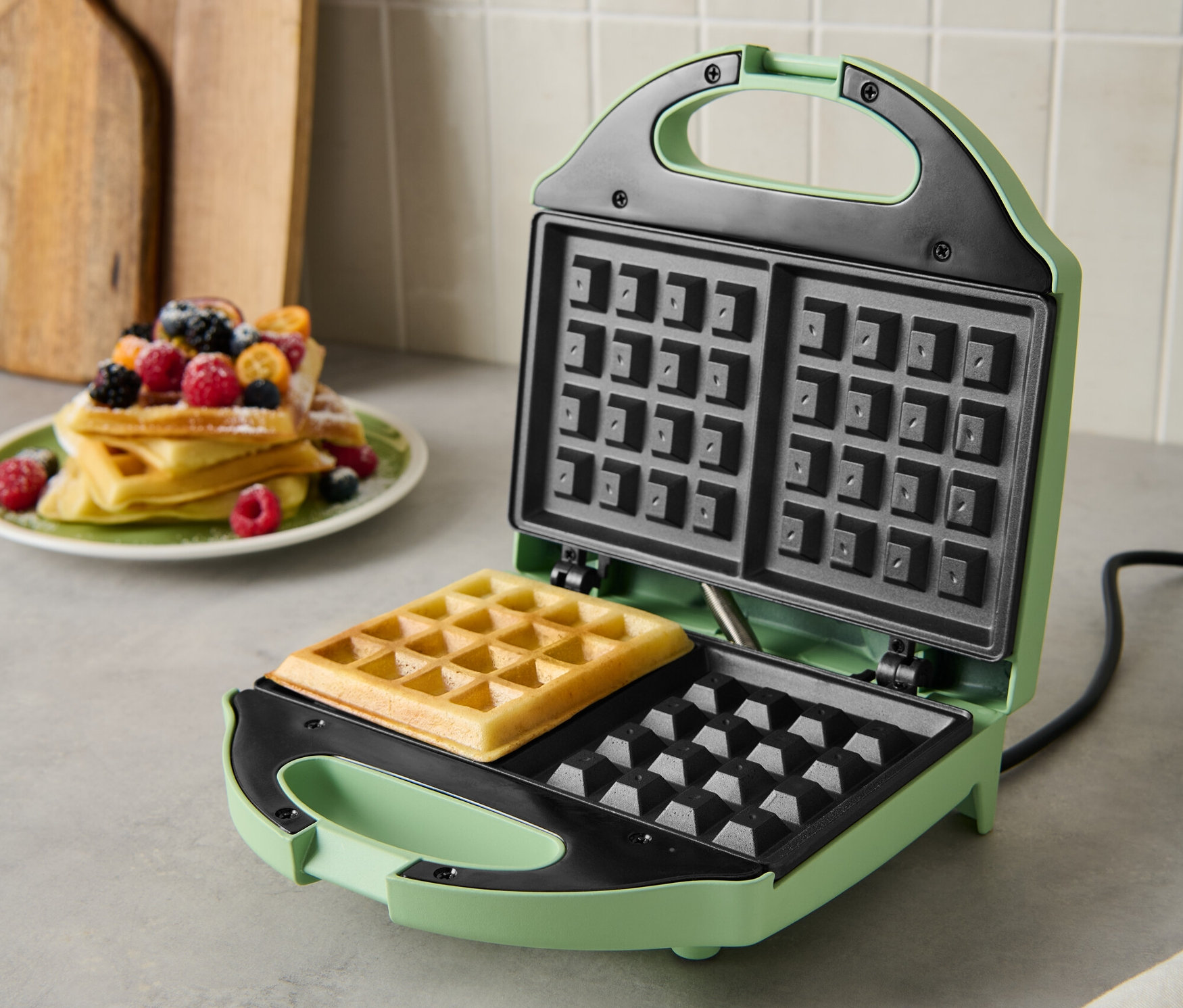 Zielona gofrownica z upieczonym gofrem w środku. Na talerzu leży stos gofrów z owocami.