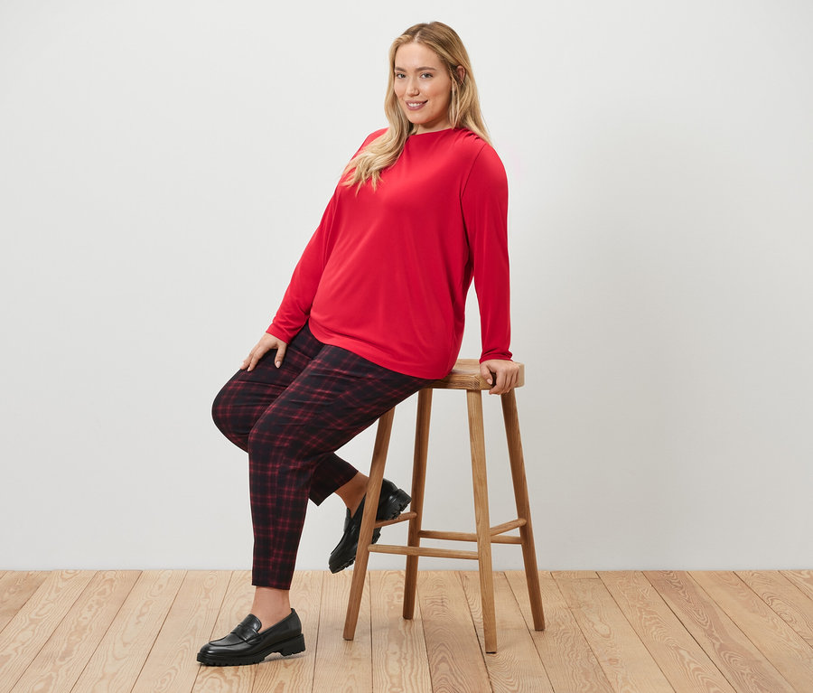 Kobieta siedzi na stołku i ma na sobie spodnie stretchowe w kratę oraz bluzkę z marszczeniem.