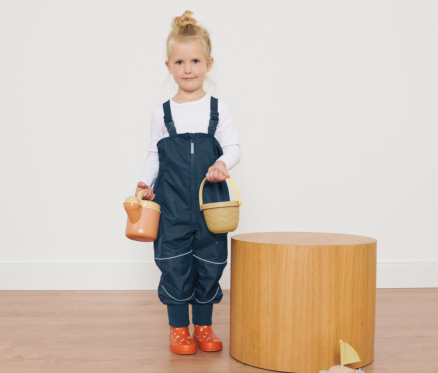 Dziewczynka w Spodnie przeciwdeszczowe trzyma zabawki obok drewnianego stolika.