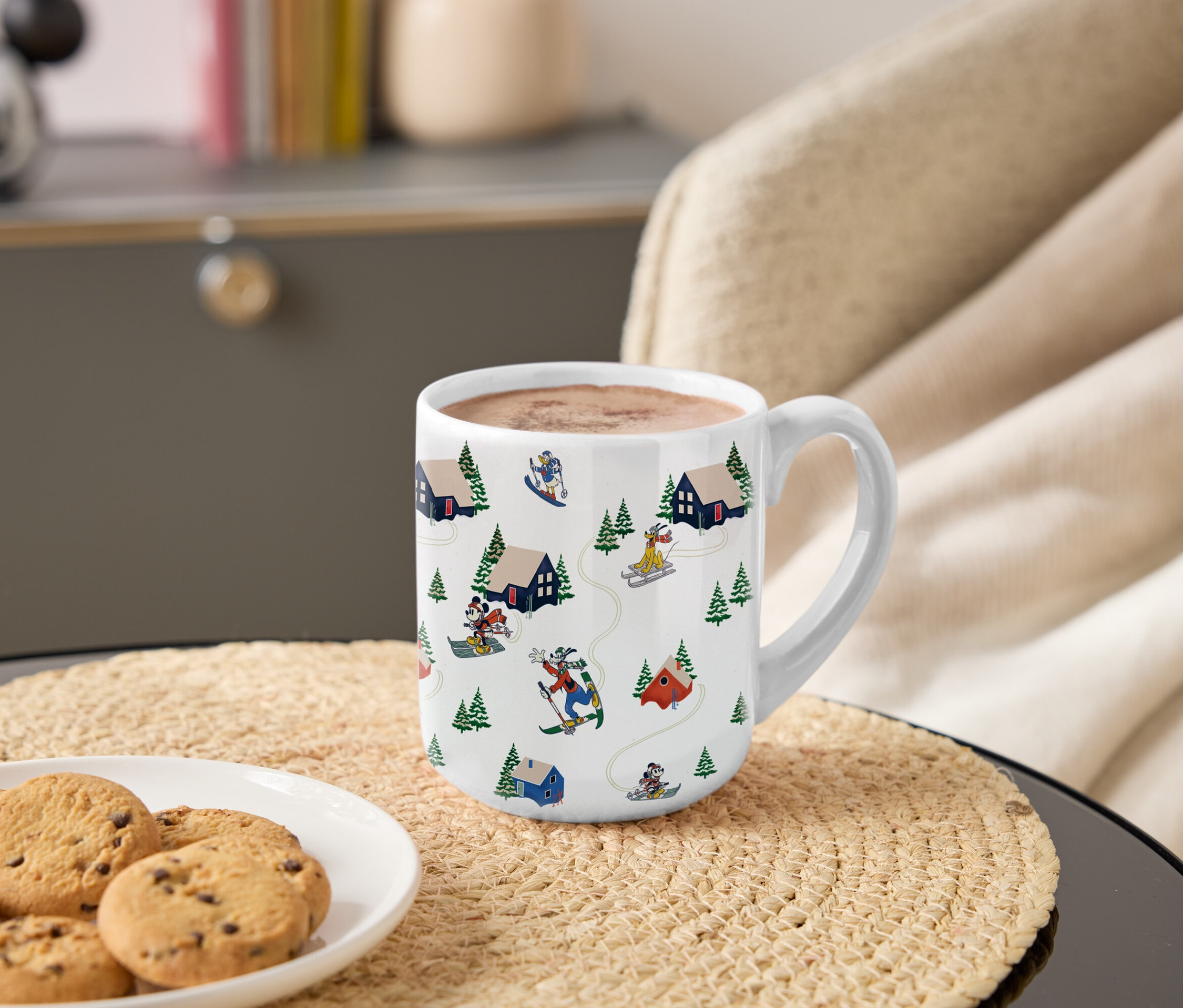 Kubek z motywem Myszki Miki i przyjaciół jeżdżących na nartach w górach stoi na stole obok talerza ciasteczek.