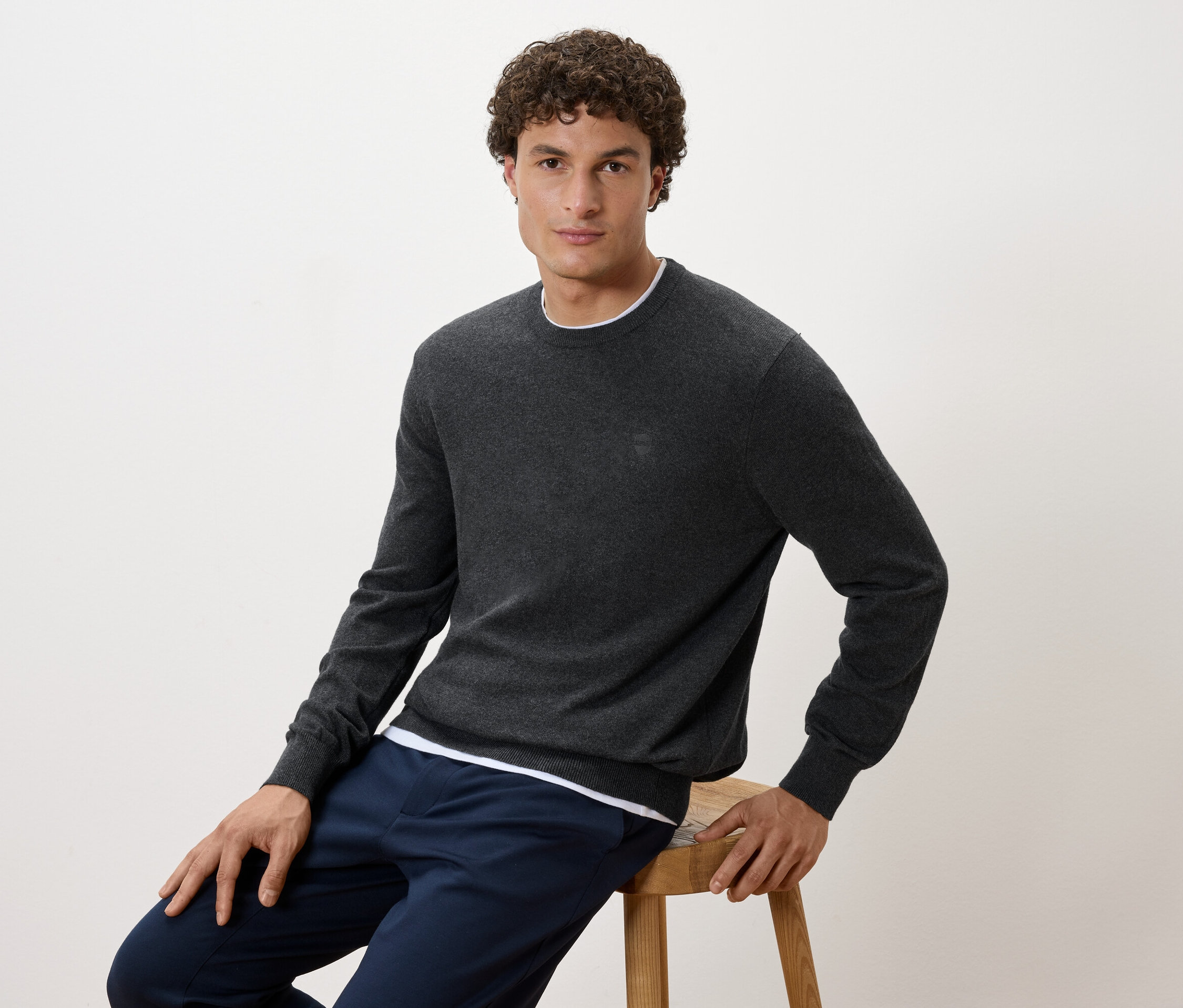 Mężczyzna siedzi na stołku, ubrany w szary sweter i granatowe spodnie.