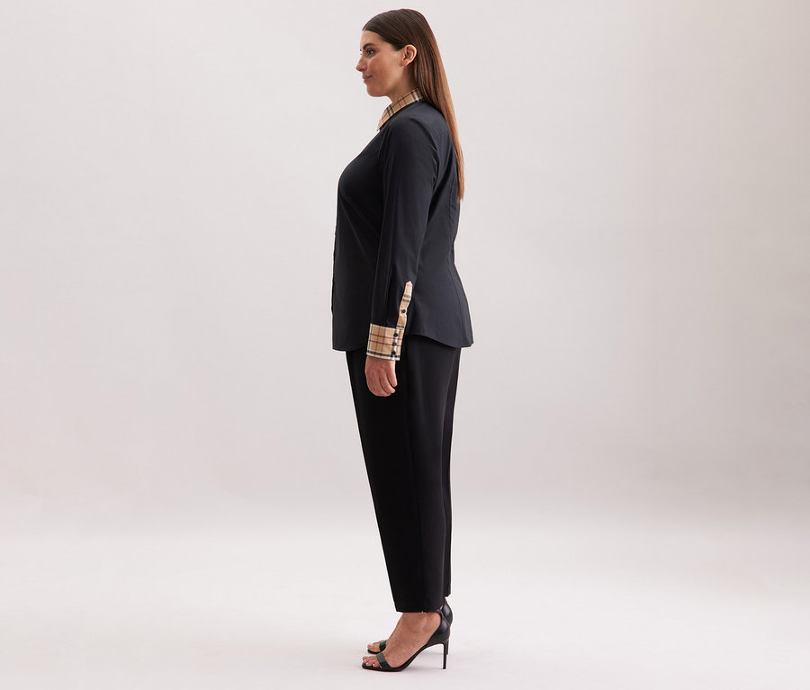 Kobieta ma na sobie czarną popelinową bluzkę Seidensticker z detalami w kratę, czarne spodnie i czarne obcasy.