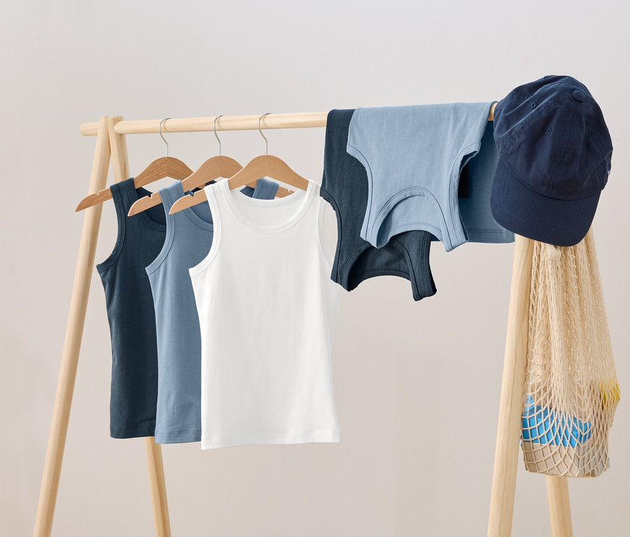 Na drewnianym wieszaku wiszą trzy podkoszulki na wieszakach, obok nich leżą dwa kolejne podkoszulki, niebieska czapka z daszkiem i siatka na zakupy.