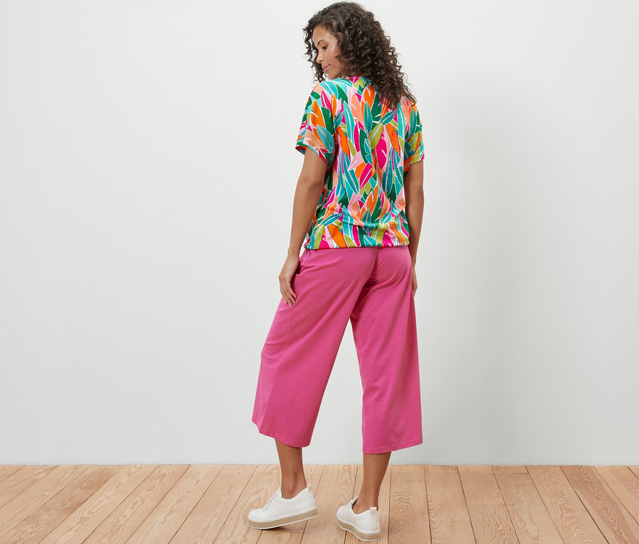 Kobieta stoi tyłem, ubrana w bluzkę z obniżoną linią ramion, ze wzorem na całej powierzchni i różowe spodnie.