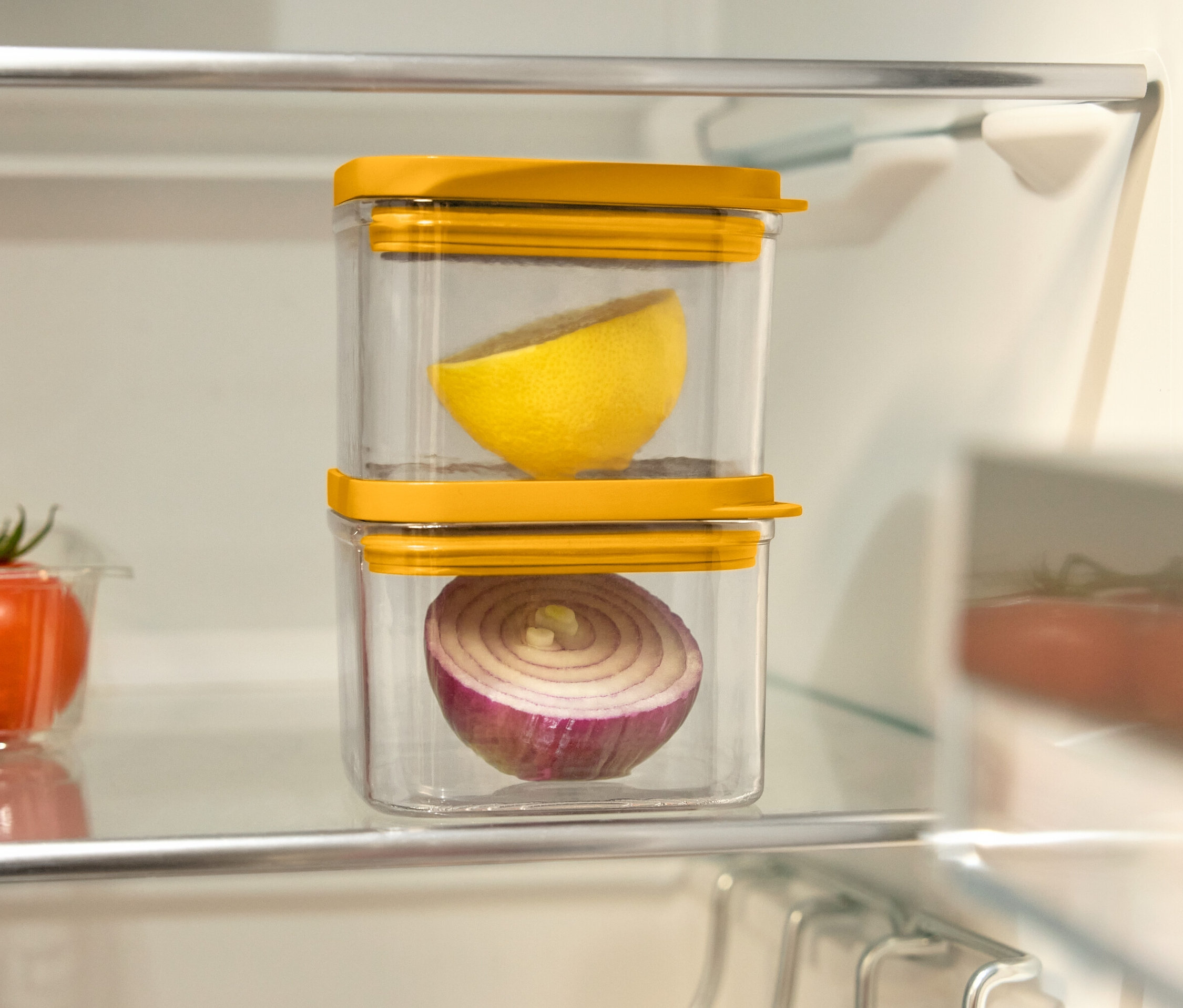 W lodówce stoją dwa przezroczyste pojemniki z żółtymi pokrywkami, jeden na drugim. W górnym pojemniku znajduje się połówka cytryny, a w dolnym połówka czerwonej cebuli. Na lewo od nich znajduje się plastikowy pojemnik z pomidorami.