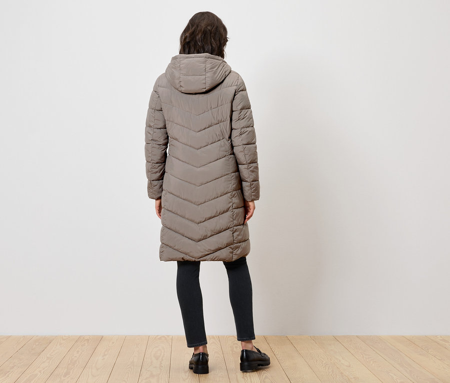 Kobieta tyłem do kamery ma na sobie długi pikowany płaszcz z kapturem, czarne spodnie i czarne buty.