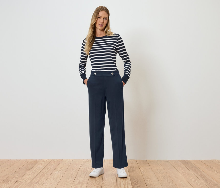 Kobieta stoi z rękami w kieszeniach. Ma na sobie bluzkę w paski z długim rękawem i granatowe spodnie.