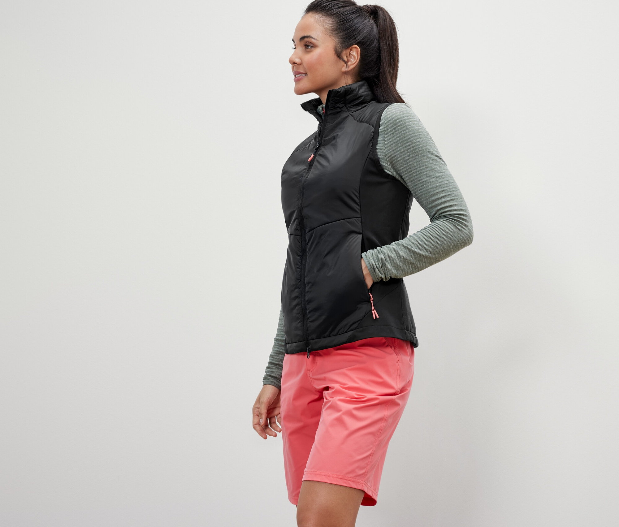 Kobieta pozuje w kamizelce hybrydowej „pack light”, oliwkowej bluzce i różowych szortach.