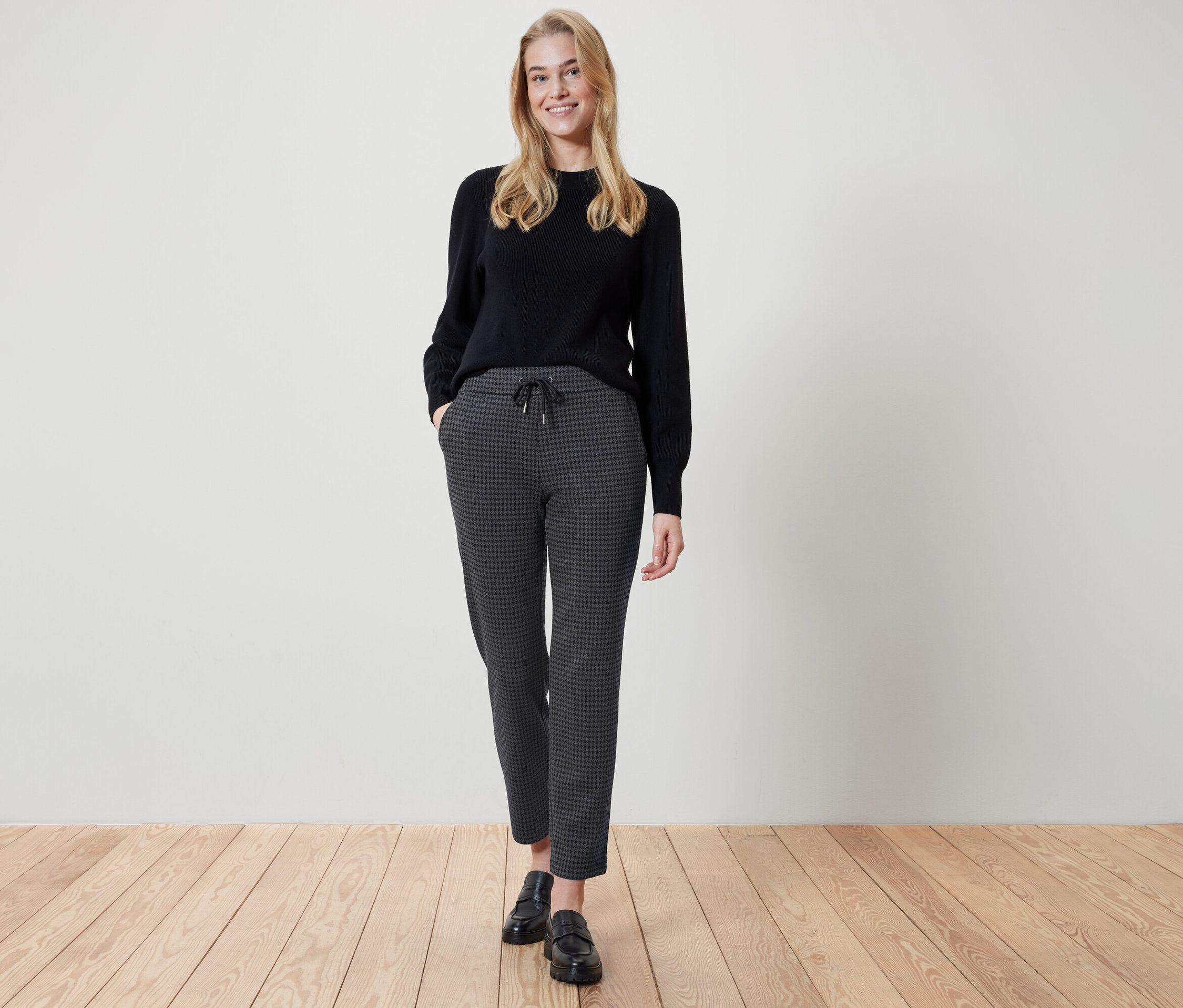 Kobieta stoi z rękami w kieszeniach spodni w kratę. Ma na sobie czarny sweter i czarne buty.