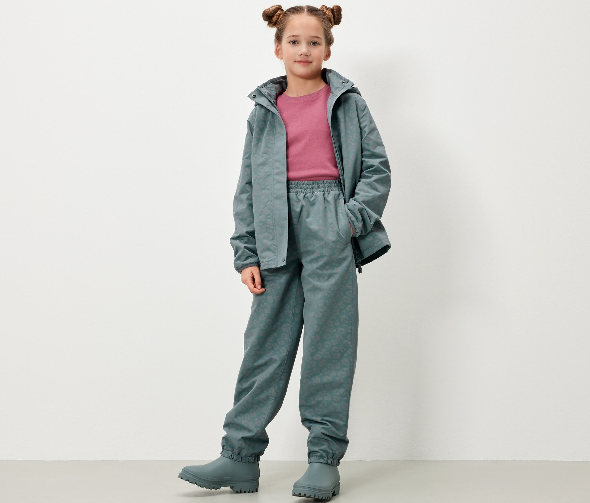 Dziewczynka stoi i pozuje w dziecięcych spodniach przeciwdeszczowych z elementami odblaskowymi, kurtce i kaloszach w kolorze szarozielonym.