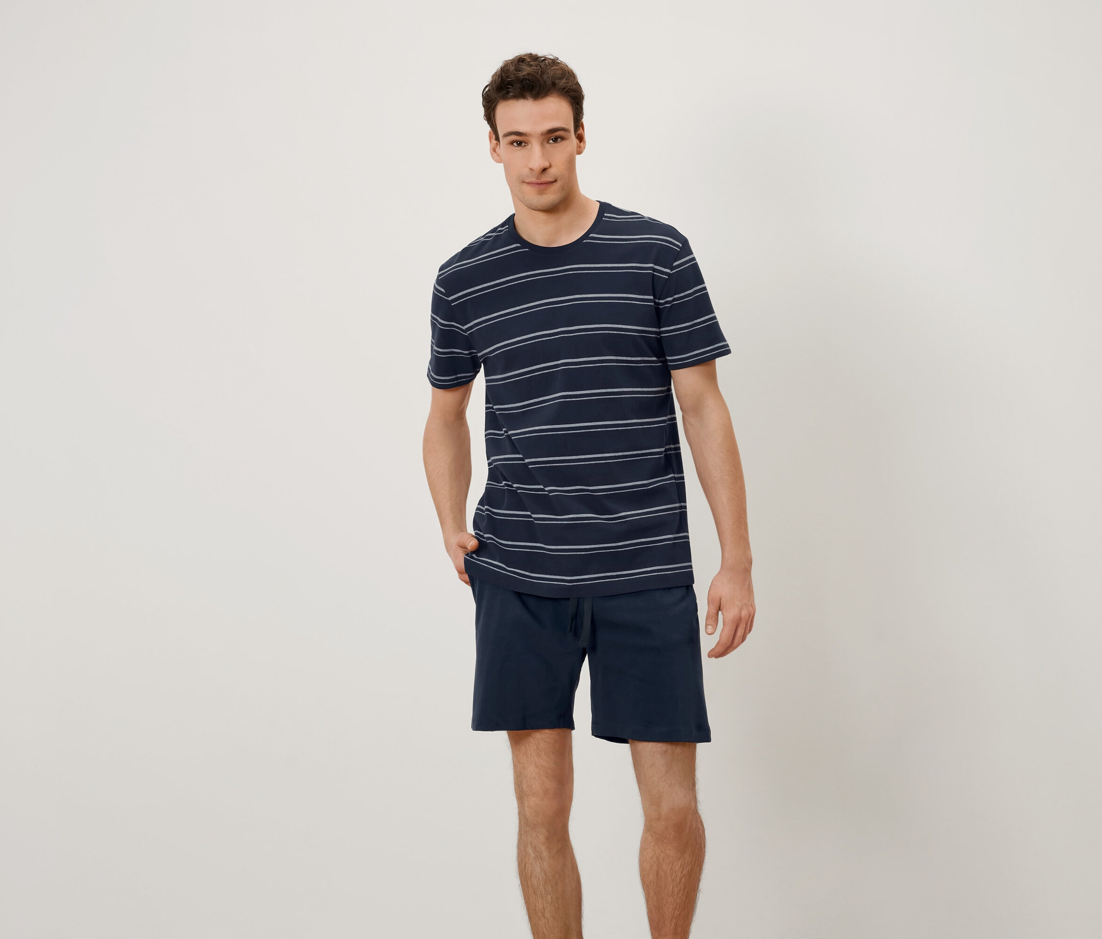 Mężczyzna stoi z rękami w kieszeniach. Ma na sobie koszulkę w poziome paski i szorty.