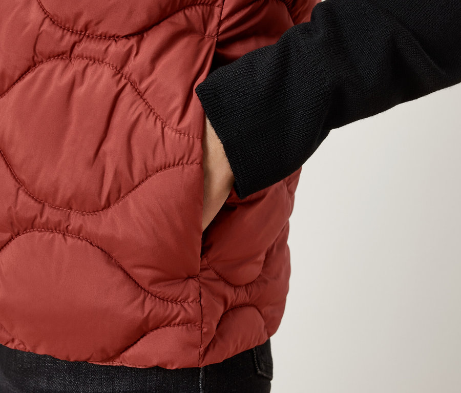 Zbliżenie na osobę w brązowej pikowanej kamizelce z ręką w kieszeni.