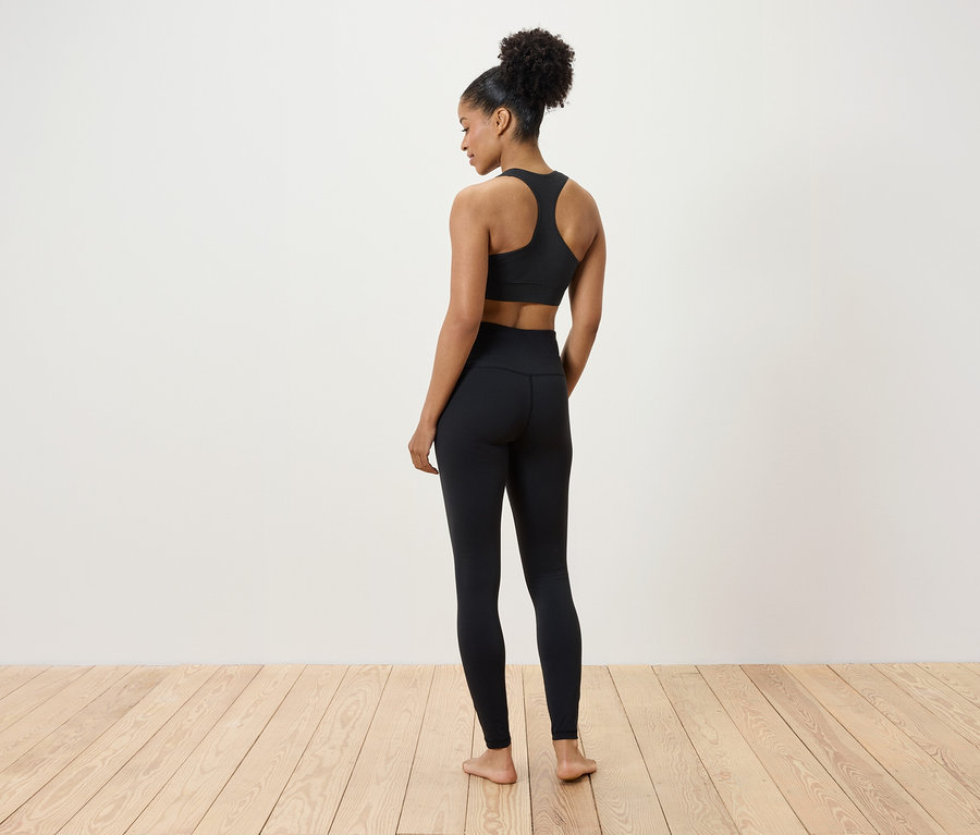 Kobieta w czarnym biustonoszu sportowym i legginsach stoi na drewnianej podłodze, tyłem do kamery.