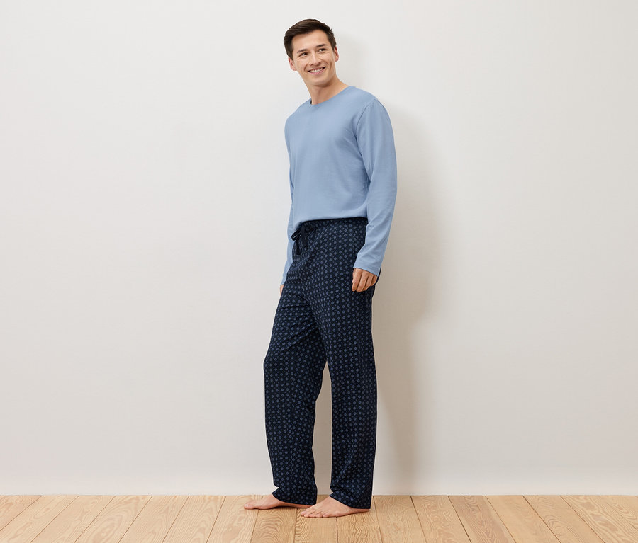 Mężczyzna stoi boso na drewnianej podłodze na tle białej ściany, ubrany w jasnoniebieski sweter i granatowe spodnie od piżamy w kropki.