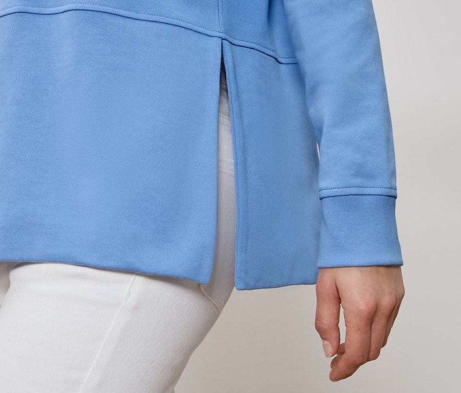 Osoba nosi błękitną bluzę dresową z rozcięciem bocznym i białe spodnie.