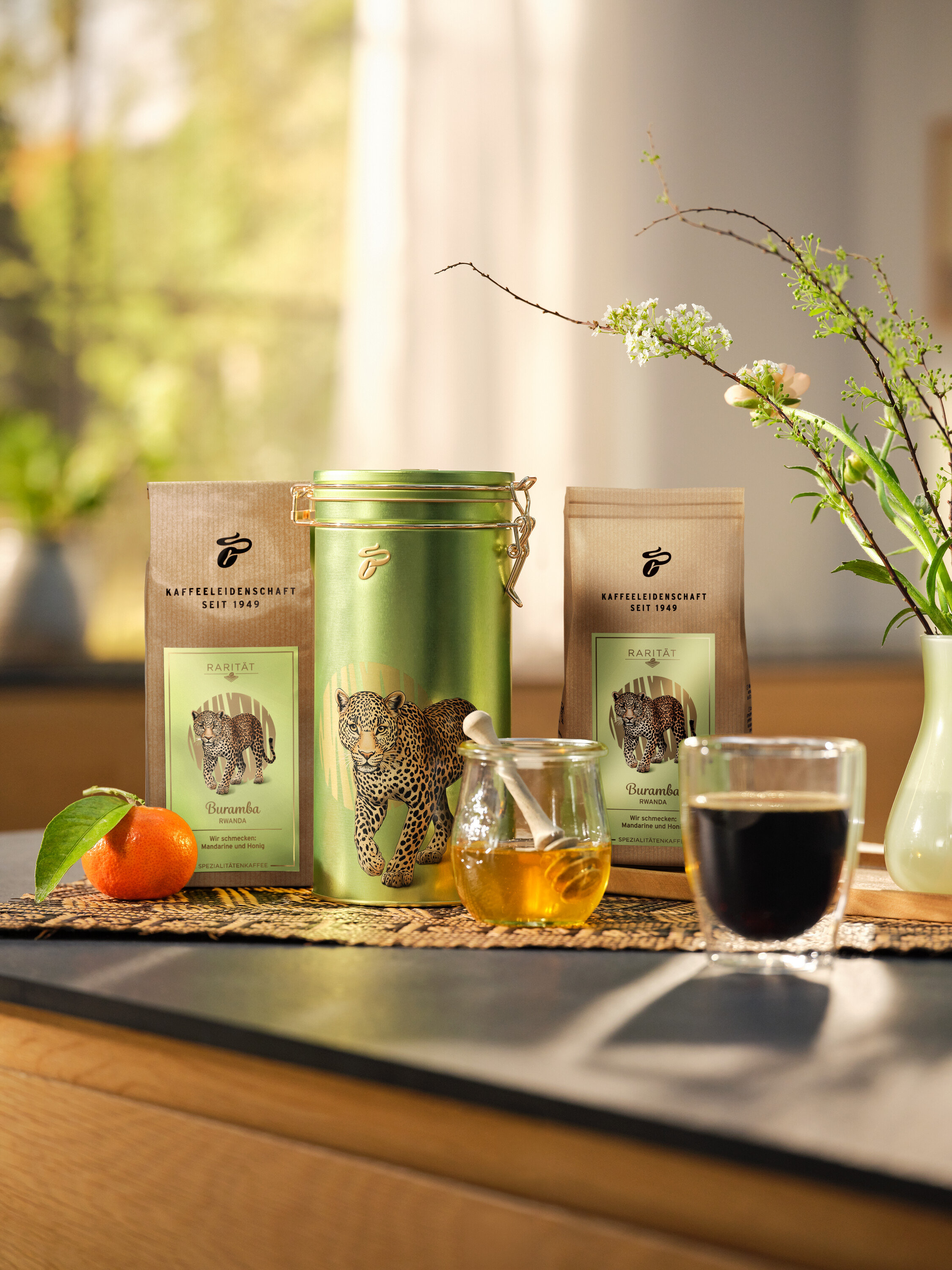 Na stole stoją dwie paczki kawy, zielona puszka z leopardem, słoik miodu, mandarynka i filiżanka kawy.