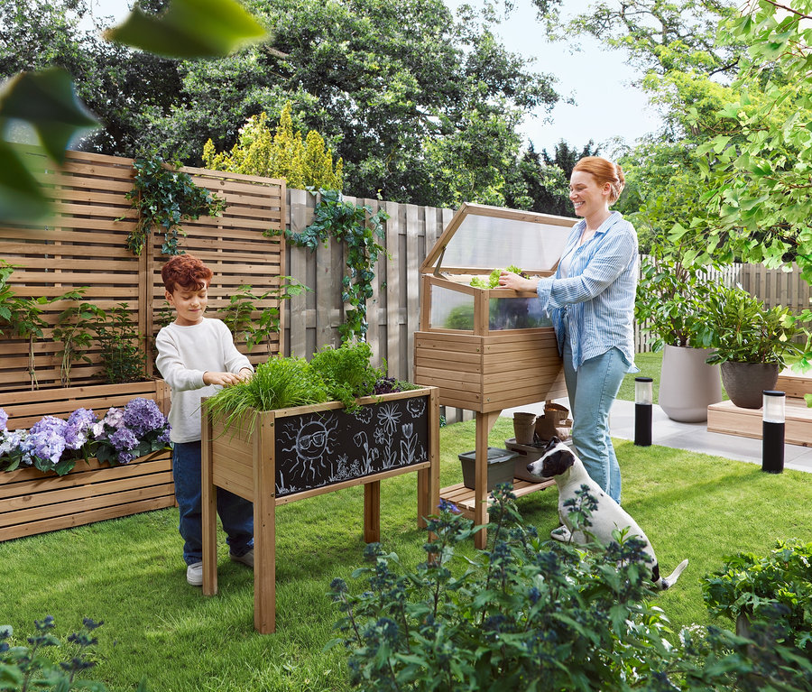 Kobieta otwiera inspekt, podczas gdy dziecko stoi obok podwyższonej grządki. Pies siedzi na trawie.