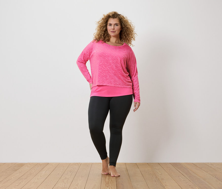 Kobieta o kręconych włosach w różowej bluzce z długim rękawem w zebrę i czarnych legginsach.