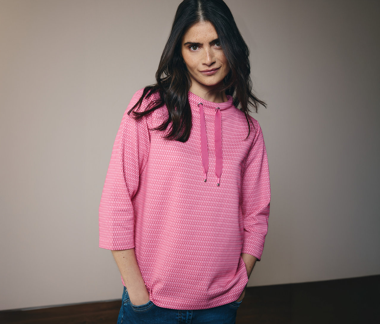 Kobieta w dżinsach i różowej koszulce CECIL z kapturem i sznurkiem. Ręce ma w kieszeniach spodni.