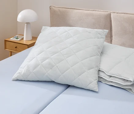 Na jasnoniebieskim łóżku leży biała, pikowana poduszka Cell Solution® (90 x 70 cm). Obok znajdują się inne poduszki Cell Solution® (80 x 80 cm, 100 x 65 cm).