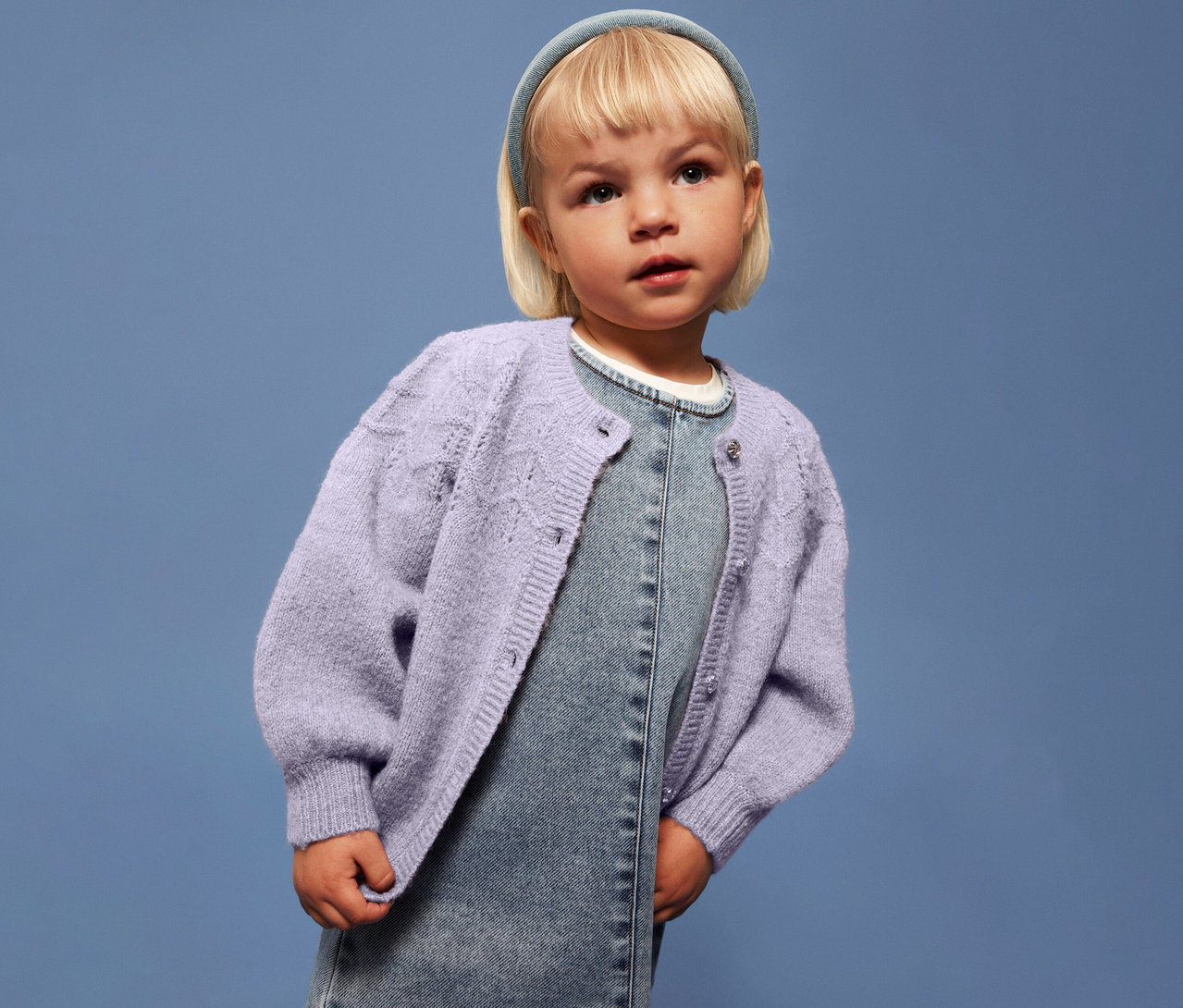 Dziewczynka z blond włosami ma na sobie jasnofioletowy kardigan dziecięcy NAME IT na dżinsowej sukience na niebieskim tle.
