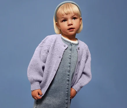 Dziewczynka z blond włosami ma na sobie jasnofioletowy kardigan dziecięcy NAME IT na dżinsowej sukience na niebieskim tle.