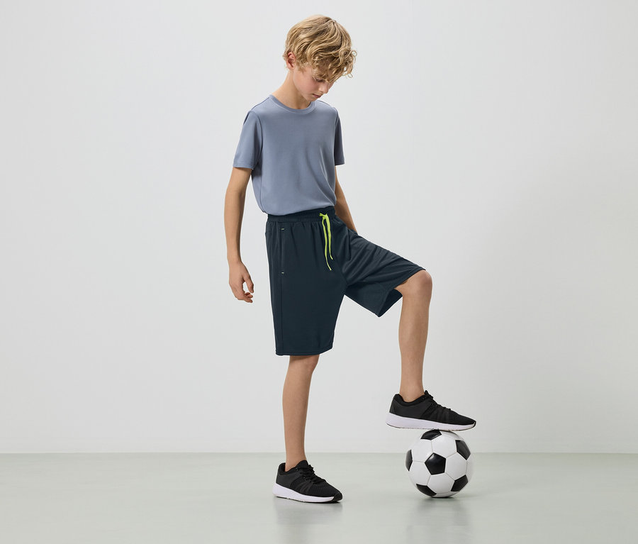 Blond chłopiec w szarej koszulce i ciemnych szortach ze stopą na piłce nożnej.