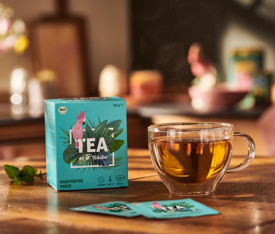 Opakowanie BIO TEA by Tchibo Mięta obok filiżanki herbaty i liści mięty na drewnianym stole.