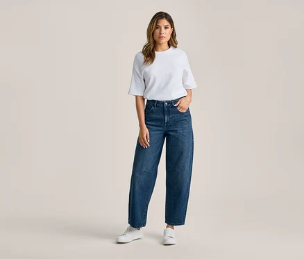 Kobieta stoi, ubrana w biały T-shirt, ciemnoniebieskie szerokie jeansy i białe sneakersy. Jej prawa ręka jest w kieszeni spodni.
