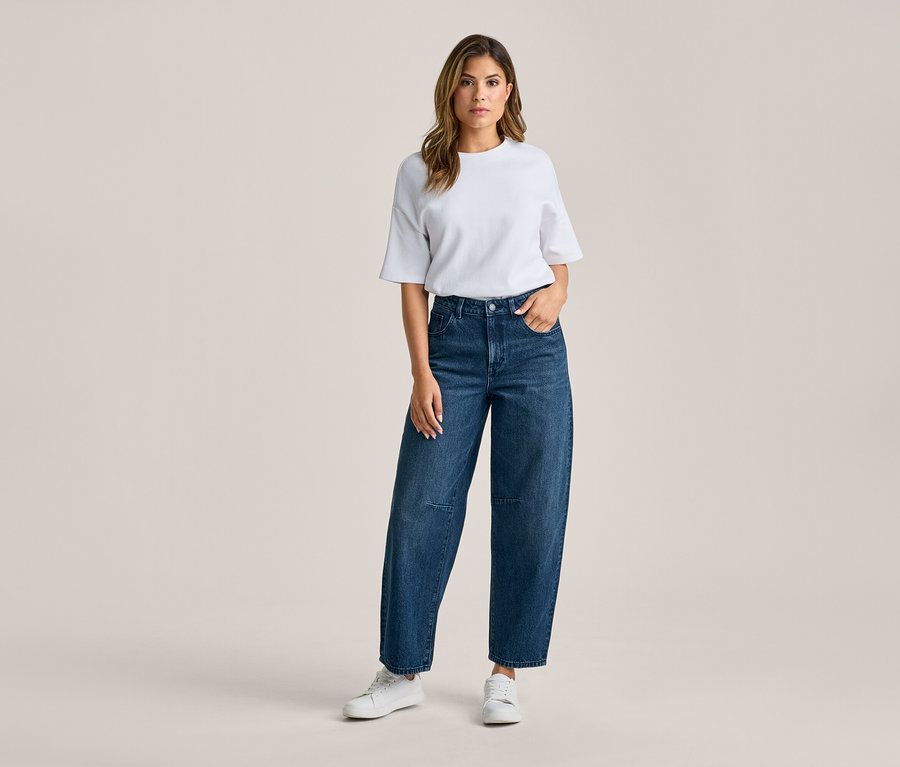 Kobieta stoi, ubrana w biały T-shirt, ciemnoniebieskie szerokie jeansy i białe sneakersy. Jej prawa ręka jest w kieszeni spodni.