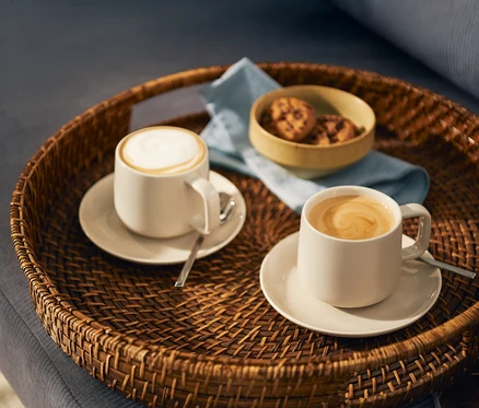 Dwie jasne filiżanki kawy, jedna z mleczną pianką, a druga z wzorem latte art, stoją na pasujących spodkach z łyżeczkami na okrągłej plecionej tacy. Na tacy znajduje się również mała miseczka z ciasteczkami i jasnoniebieska serwetka.