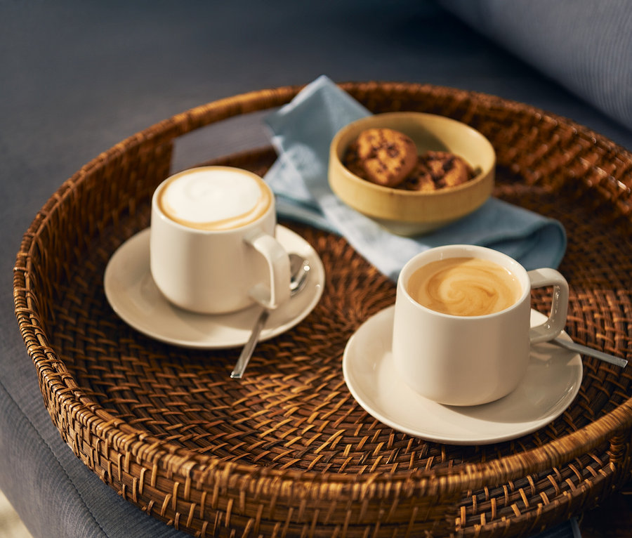 Dwie jasne filiżanki kawy, jedna z mleczną pianką, a druga z wzorem latte art, stoją na pasujących spodkach z łyżeczkami na okrągłej plecionej tacy. Na tacy znajduje się również mała miseczka z ciasteczkami i jasnoniebieska serwetka.