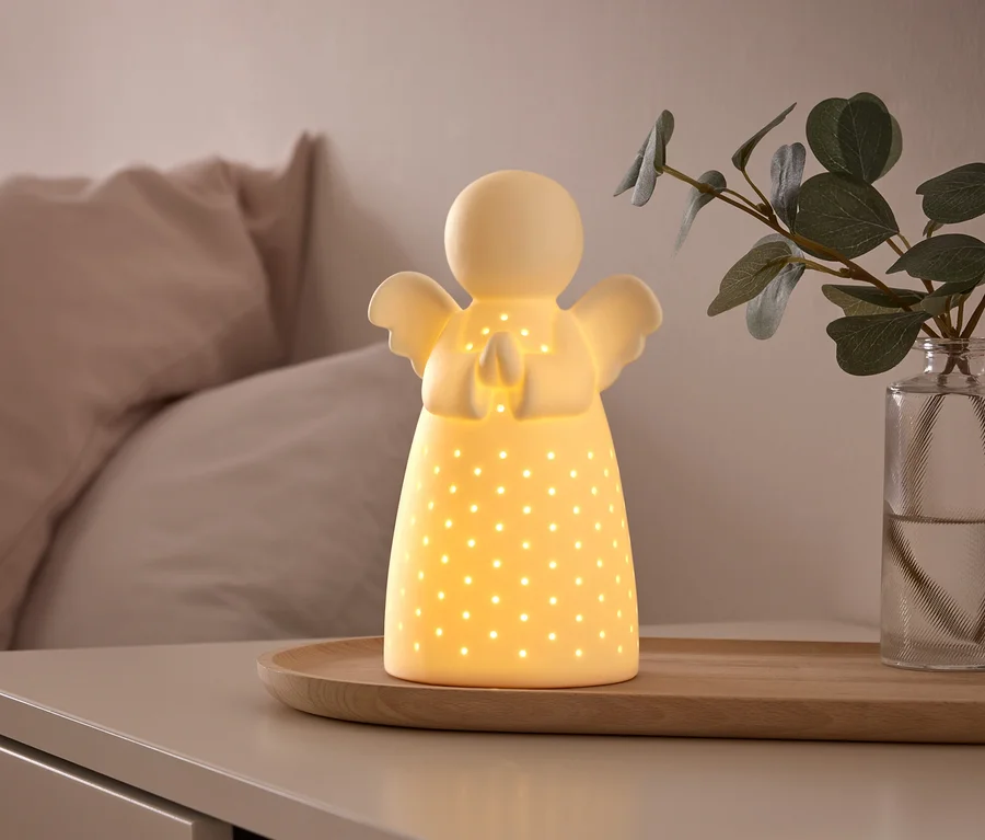 Świecąca, biała ceramiczna lampka w kształcie anioła z małymi perforacjami stoi na drewnianej tacy na stoliku nocnym, obok wazonu z zieloną rośliną. W tle widać łóżko z jasnoróżowymi poduszkami.