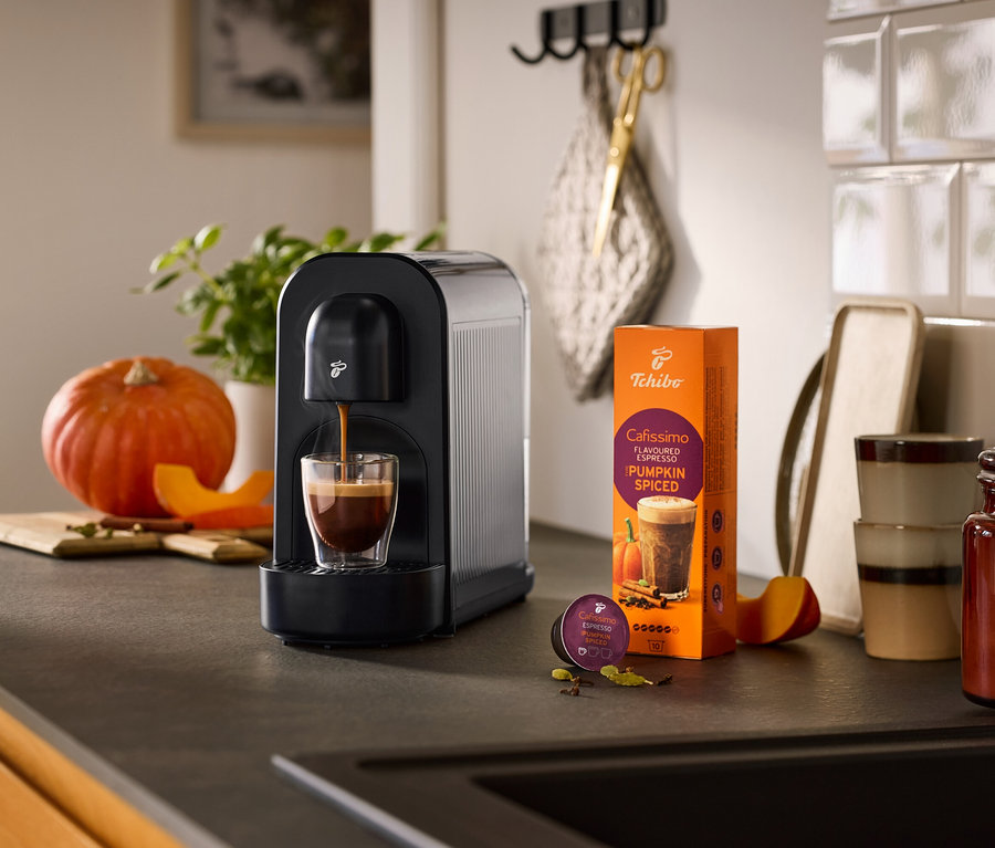 Czarny ekspres do kawy Cafissimo now parzy espresso do szklanki, obok opakowanie kawy Cafissimo Pumpkin Spiced i kapsułka, otoczone dynią i przyprawami na blacie kuchennym.