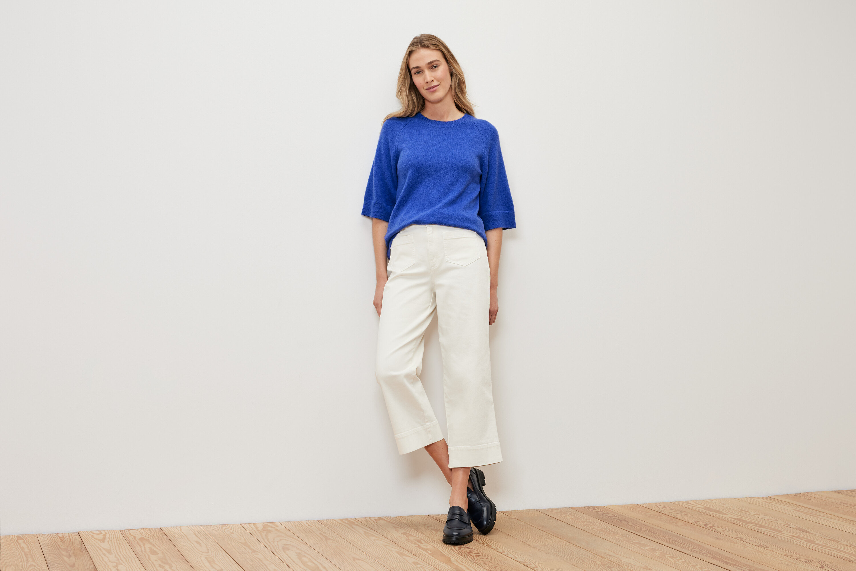 Kobieta stoi na tle białej ściany, ubrana w kobaltowy sweter, białe spodnie i czarne buty.