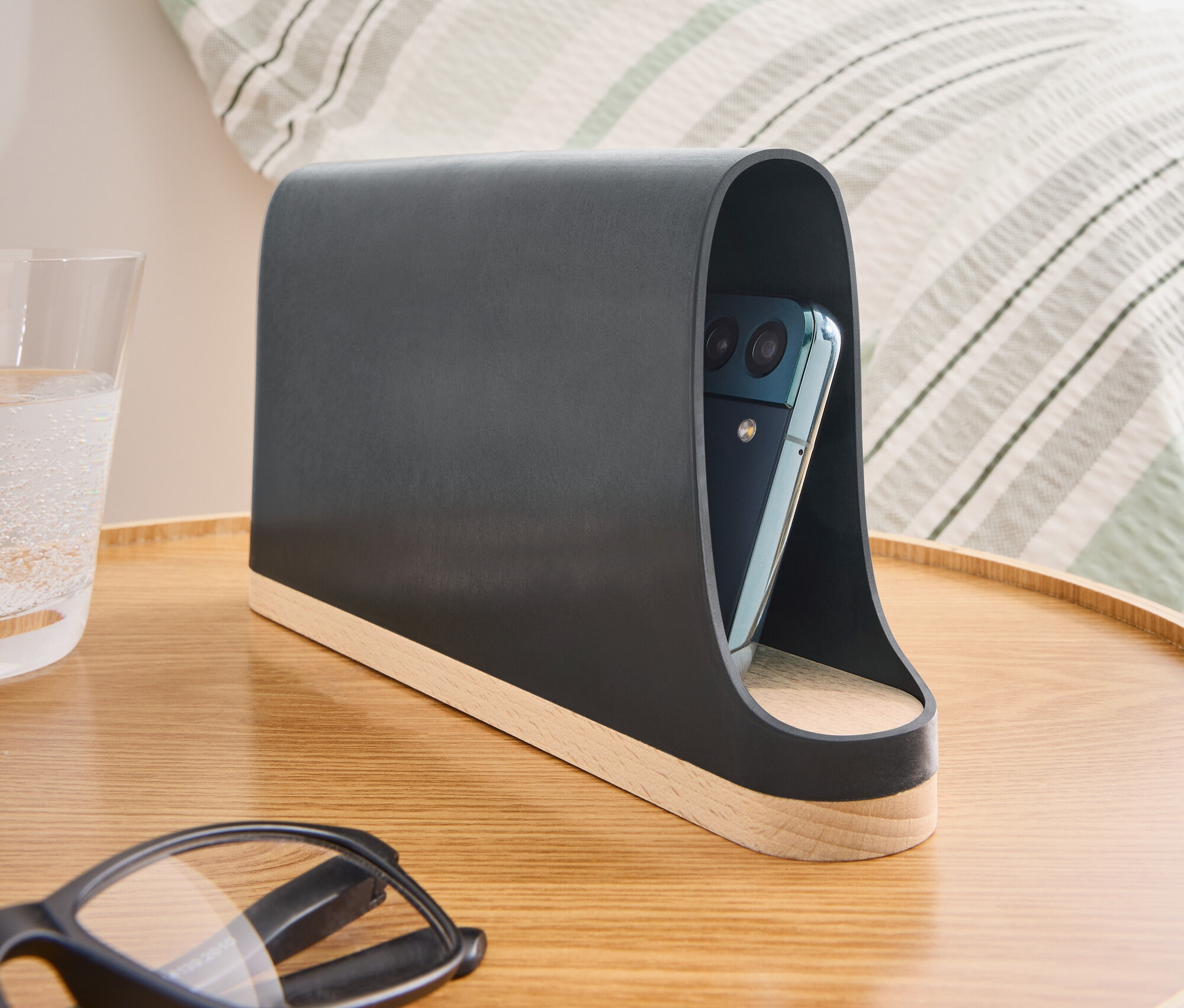 Na stoliku nocnym stoi ciemnoszary uchwyt na telefon z drewnianą podstawą i telefonem w środku.