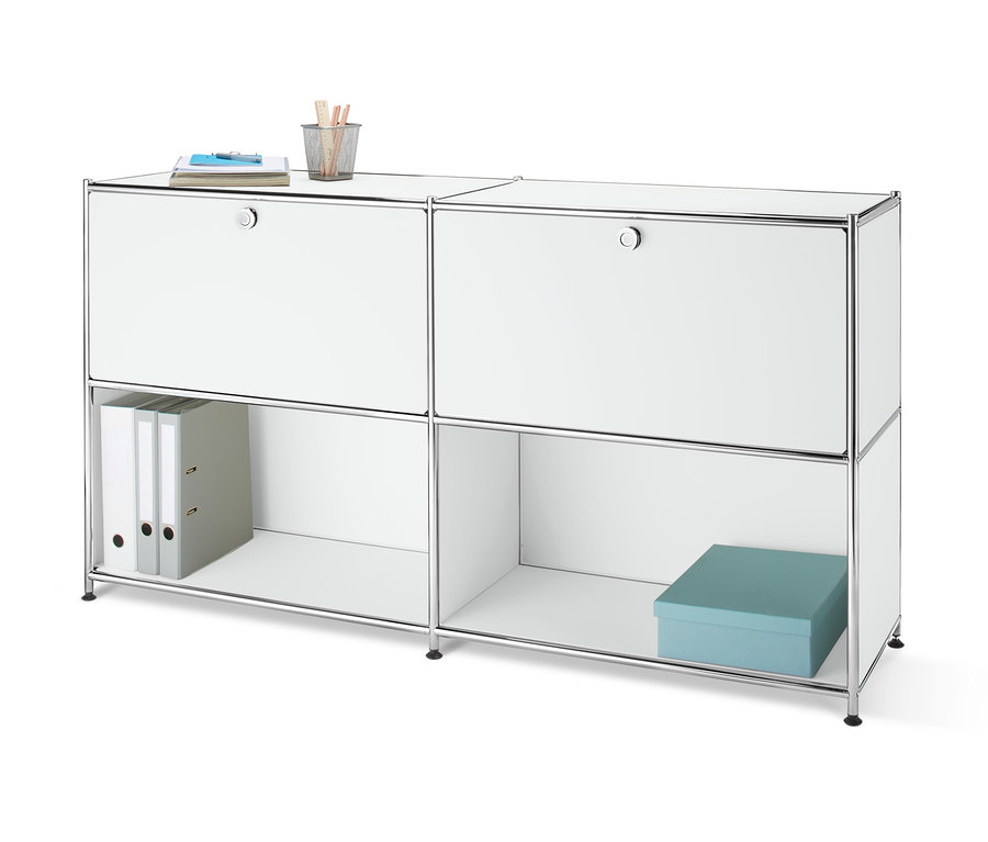 Widok z przodu metalowej szafki typu sideboard z 2 przegrodami z klapami, montowanymi w dowolnym miejscu, na której leży notatnik i szklanka z długopisami. W otwartej półce znajdują się dwa segregatory i niebieskie pudełko.