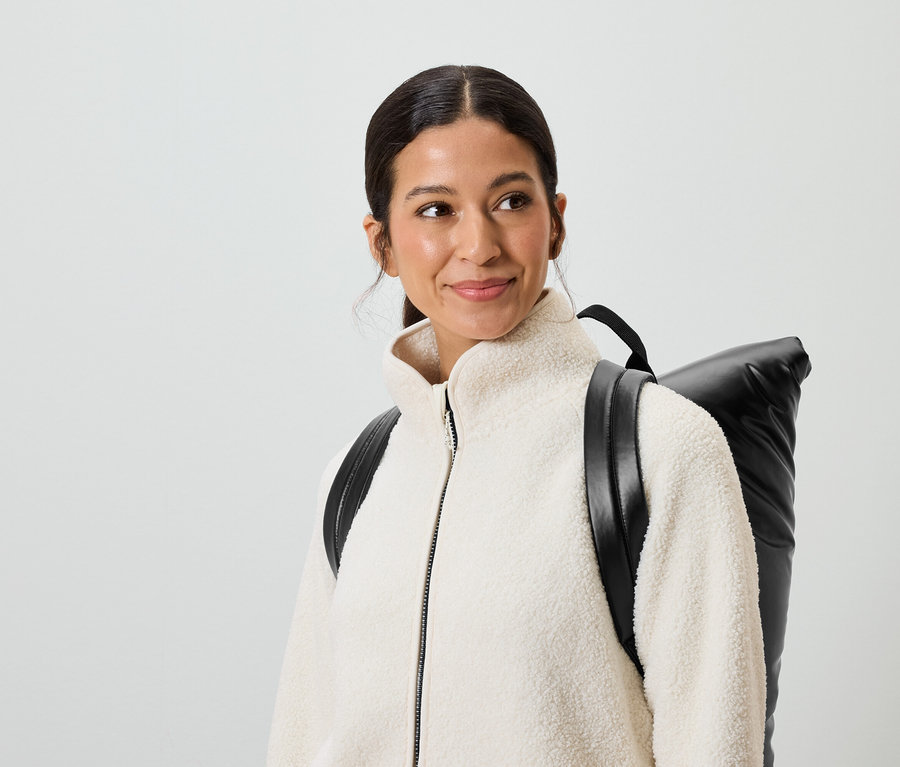 Uśmiechnięta kobieta z brązowymi włosami i brązowymi oczami ma na sobie kremową kurtkę i czarny plecak.