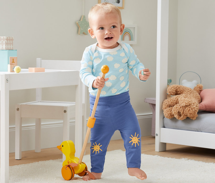 Małe dziecko bawi się żółtą kaczką na kółkach w pokoju dziecięcym, ubrane w 4-częściowy zestaw niemowlęcy z bawełny ekologicznej.