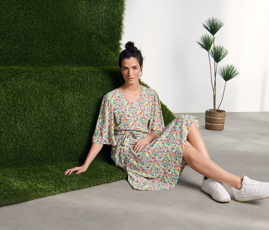 Kobieta siedzi na podłodze obok zielonej ściany, ubrana w sukienkę z nadrukiem.