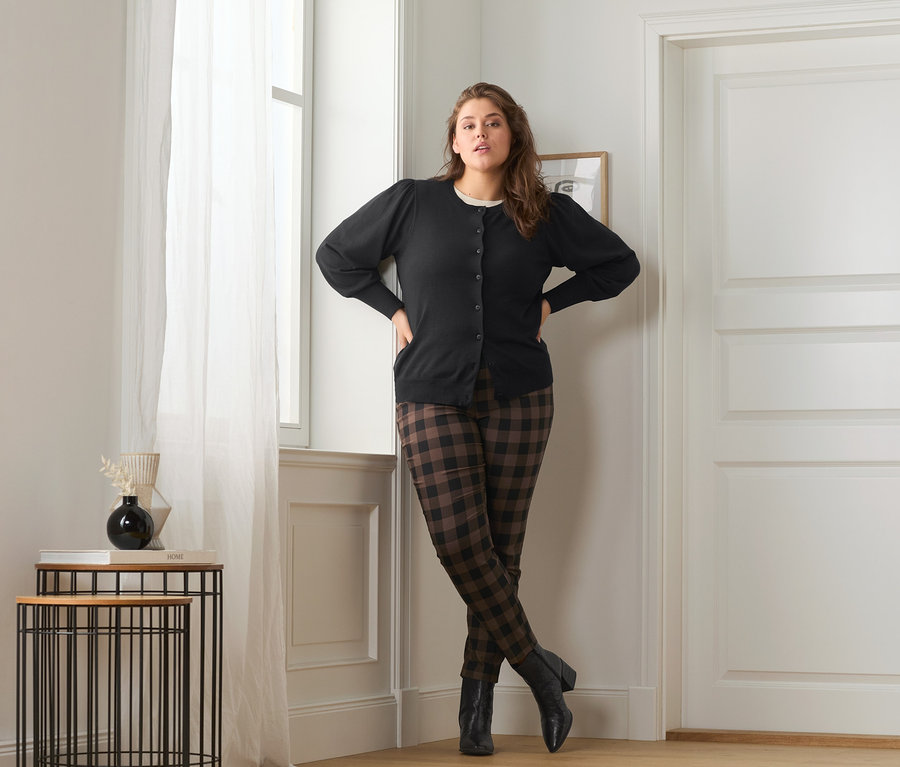 Kobieta pozuje w swetrze z delikatnej dzianiny z bufiastymi rękawami, spodniach stretchowych w kratę i skórzanych botkach.