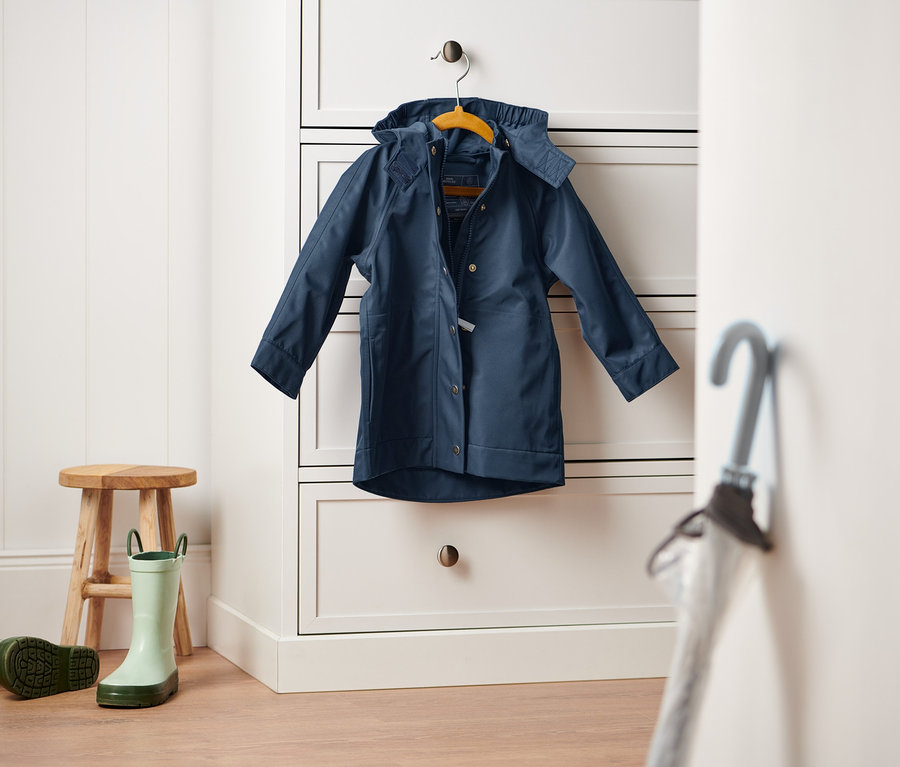 Granatowa kurtka przeciwdeszczowa wisi na białej komodzie. Na podłodze stoi drewniany stołek i para zielonych kaloszy.