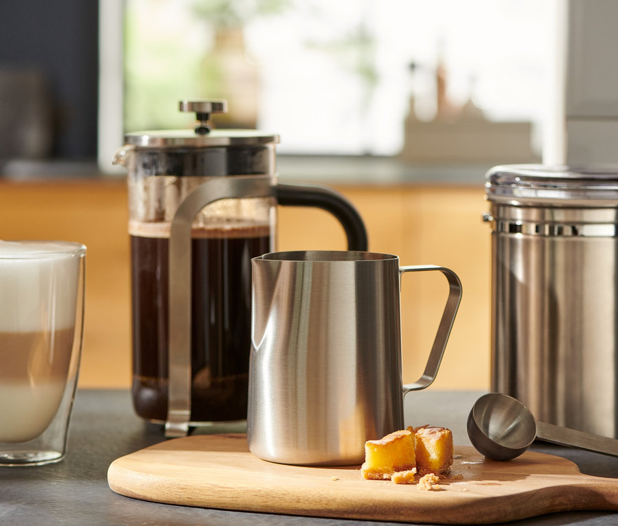 Szklanka kawy stoi obok french pressu i małego dzbanka na mleko ze stali nierdzewnej na drewnianej desce do krojenia.