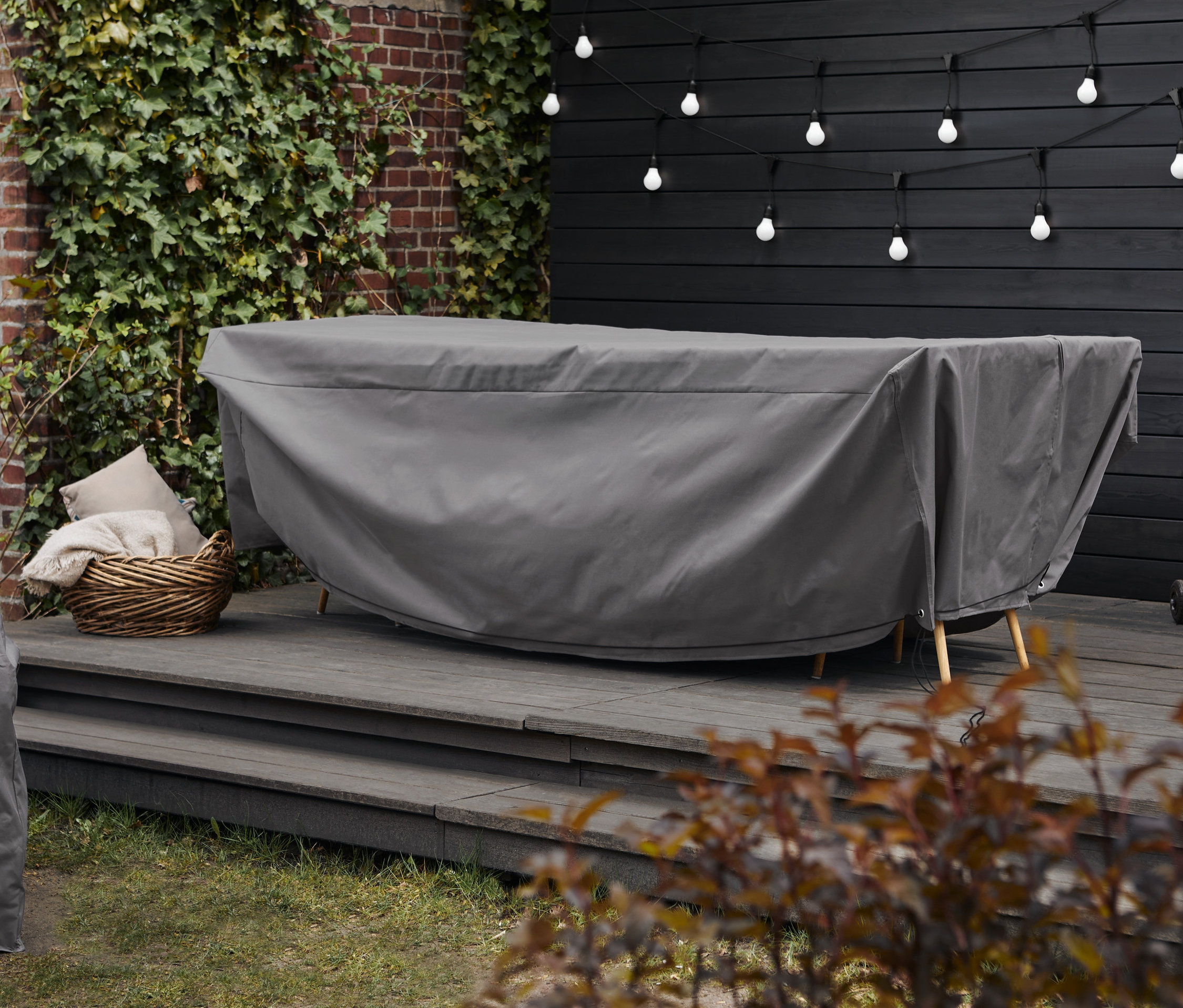 Szary pokrowiec premium zakrywa stół ogrodowy. W tle widoczna jest torba do przechowywania XXL i łańcuch świetlny LED.