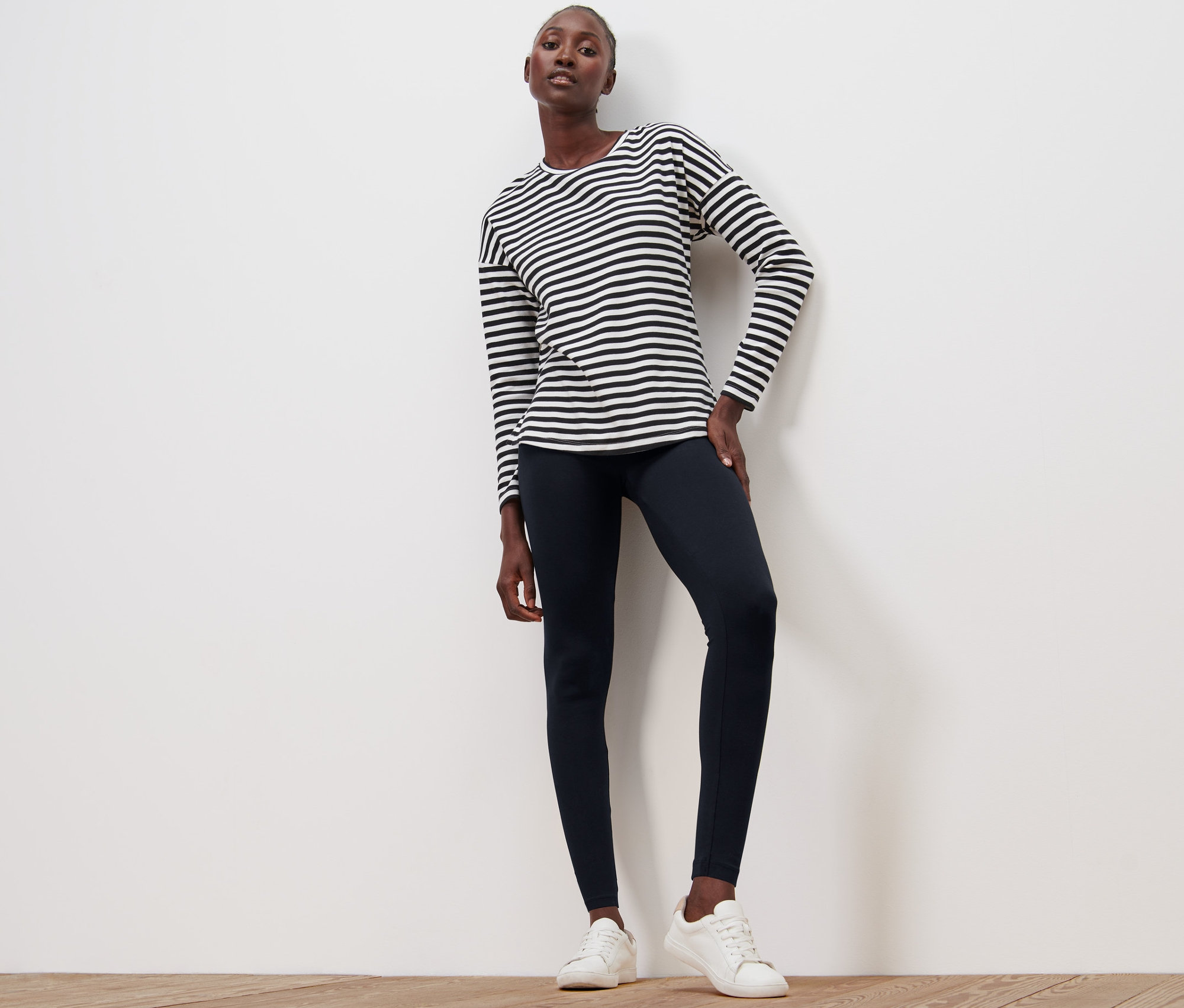 Kobieta pozuje na drewnianej podłodze w czarnych legginsach i bluzce w paski.