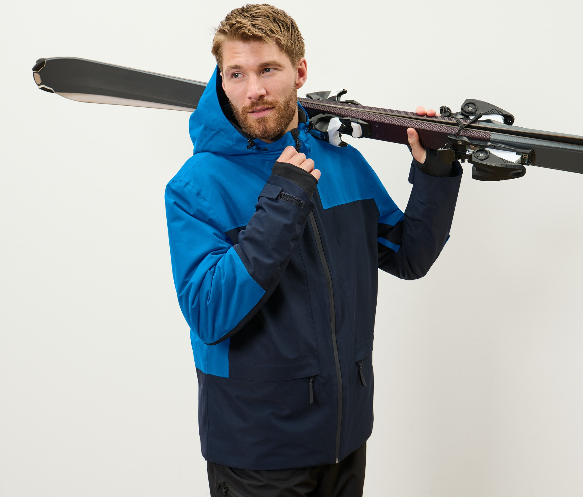 Mężczyzna w niebieskiej kurtce narciarskiej trzyma narty na ramieniu.