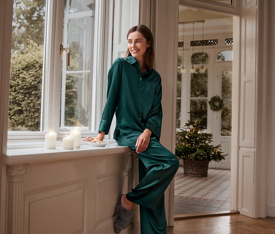 Kobieta siedzi na parapecie w zielonej jedwabnej górze i spodniach od piżamy oraz pluszowych kapciach.